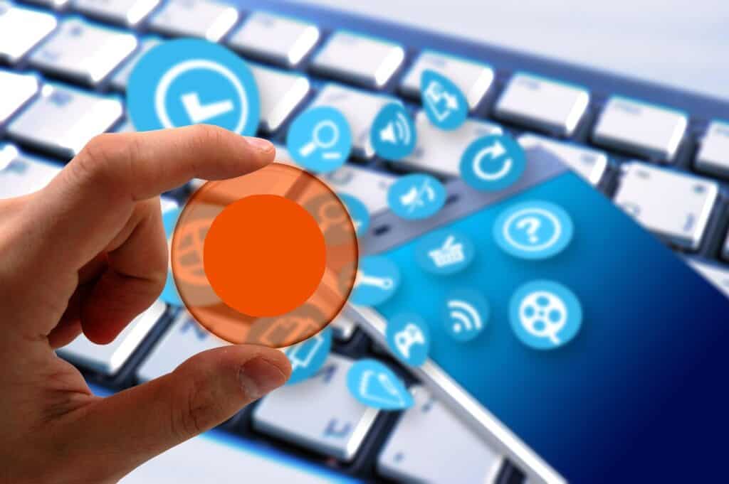 Osoba trzymająca pomarańczowy przycisk nad klawiaturą i smartfonem z ikonami aplikacji – symbol zarządzania funkcjami smart home